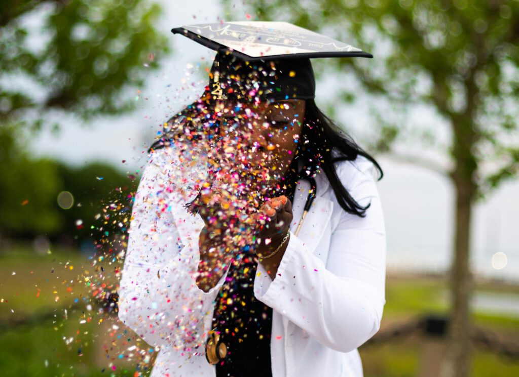 Female in graduation cap blowing confetti from hands.